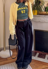 Green crop top with yellow "BRASIL 92" text, yellow cardigan, black pleated trousers, black handbag, and black shoes against a light interior.