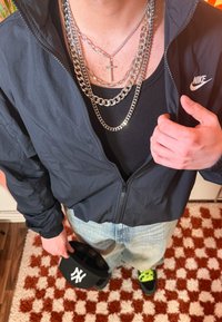 Coupe-vent noir à fermeture éclair, chaînes argentées superposées avec un pendentif en forme de croix, débardeur noir, jean bleu clair, et casquette noire avec logo blanc.