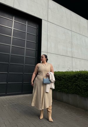 Vestido sin mangas color beige con una cintura asimétrica, combinado con botas beige hasta la rodilla y un pequeño bolso azul. Sosteniendo un suéter de color claro.