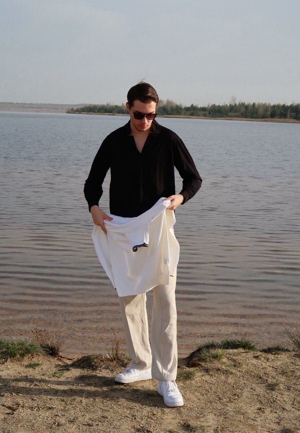 Osoba nosi crnu košulju dugih rukava i drži bijeli džemper. Ovaj outfit upotpunjuju svijetlosmeđe hlače i bijele tenisice. U pozadini se vidi voda.