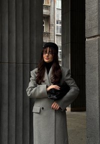 Abrigo de lana gris con diseño cruzado, mangas largas y un gran cuello. Los accesorios incluyen un bolso de mano negro y una boina negra.