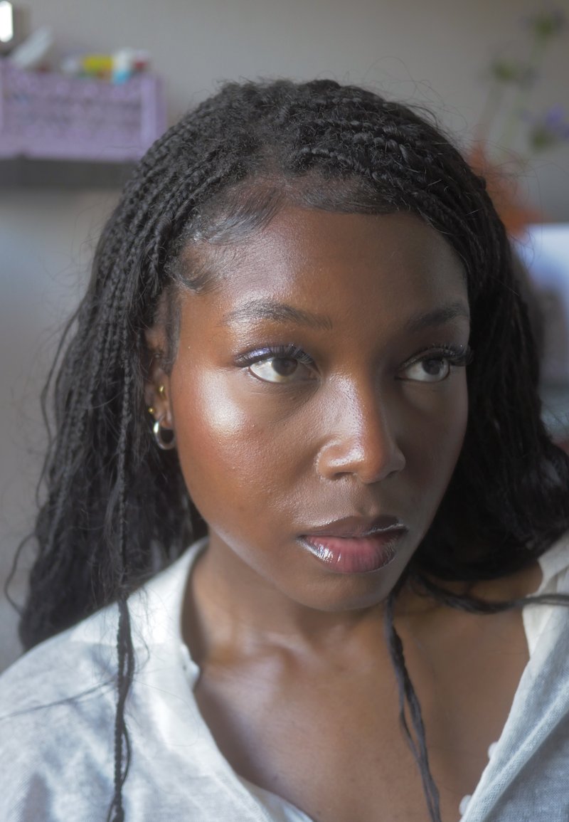 Close-up of a young person with dark skin and braided hair, wearing a light-colored garment. Features include smooth skin and subtle makeup.