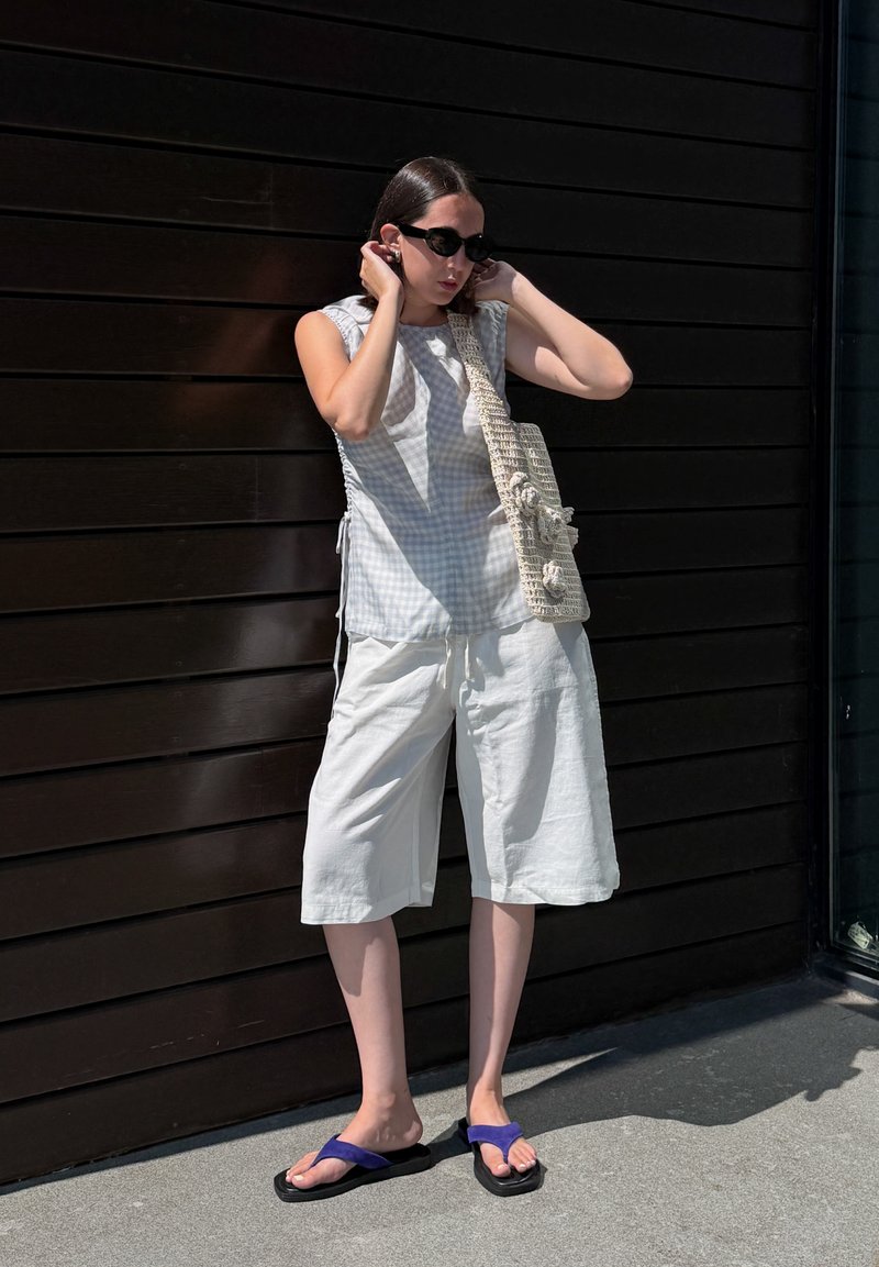 Frau stellt Sonnenbrille ein, trägt ärmelloses kariertes Top, weiße weite Shorts, lila Flip-Flops und trägt eine strukturierte Schultertasche.