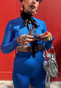 Persona in abbigliamento sportivo blu con un nastro nero al collo, anelli colorati in stile cartone animato, che tiene occhiali da sole e una borsa argentata contro un muro rosso.