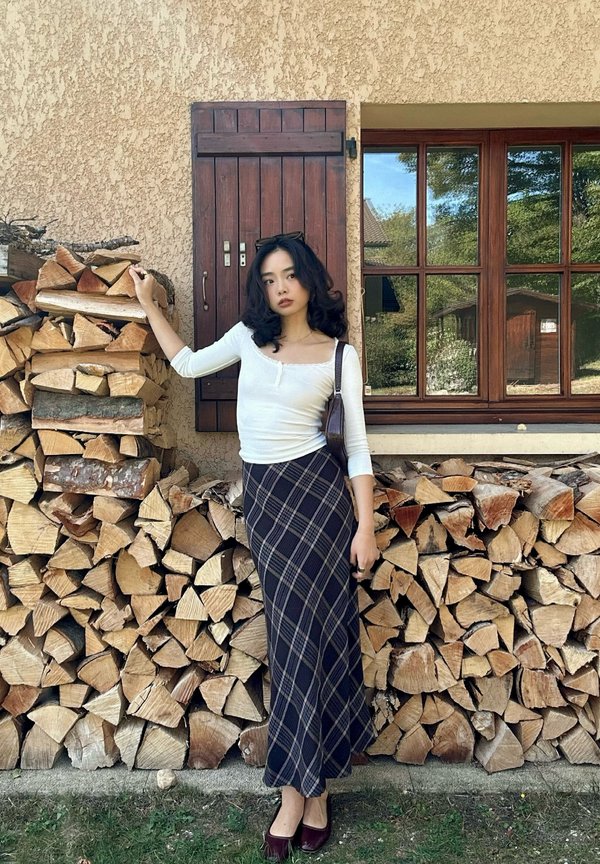 Mujer con top blanco y falda a cuadros se apoya en leña apilada frente a una casa con contraventana de madera y una ventana que refleja árboles.