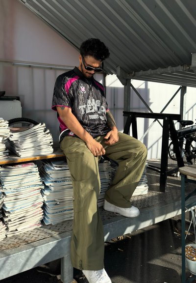 Jersey deportivo negro y rosa con el texto "MÁS TIEMPO", combinado con pantalones cargo verde claro y zapatillas blancas, sentado sobre una pila de revistas.