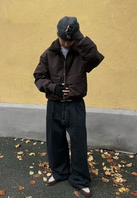 Smeđa jakna na patentni zatvarač s flis okovratnikom, široke crne traperice, siva pletena kapa i crne rukavice bez prstiju. Lišće na tlu.