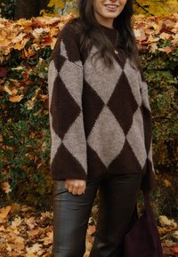 Femme portant un pull à losanges marron et beige et un pantalon en cuir noir, debout devant des feuilles d'automne et de la verdure, tenant un sac foncé.