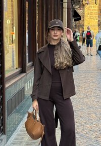 Hnedá skrátená bunda cez čierny top, spárovaná s čiernymi širokými nohavicami. Drží malú hnedú kabelku, na hlave má tmavú čiapku. Pozadie tvorí ulica vydláždená kameňmi.