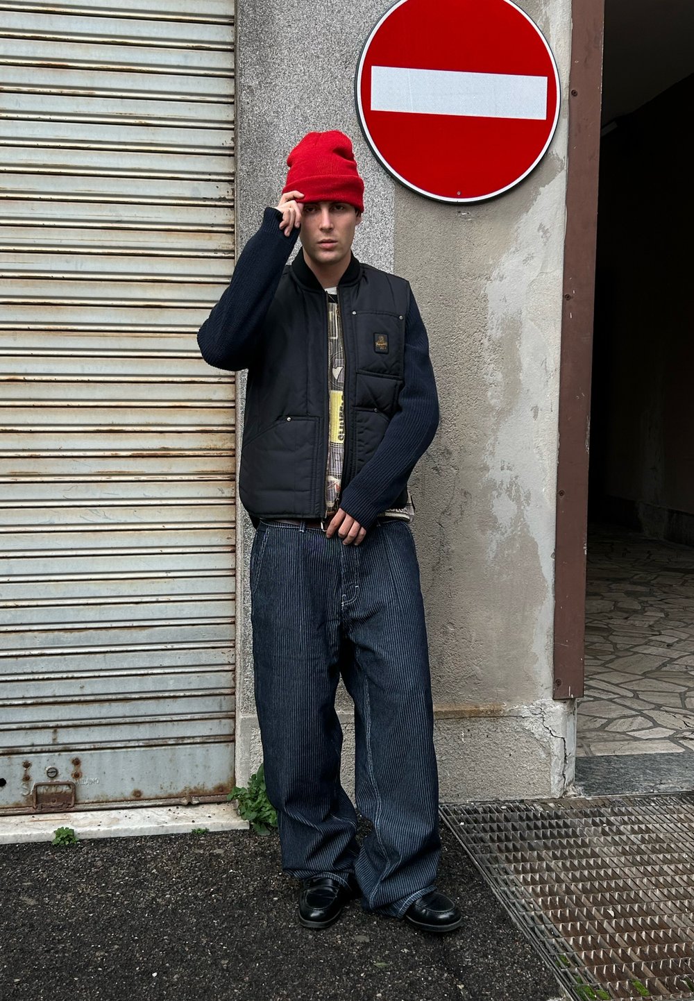 Un homme portant un bonnet rouge, un gilet noir, un pantalon large rayé et des chaussures noires se tient près d'un mur surmonté d'un panneau de signalisation d'interdiction d'entrée.