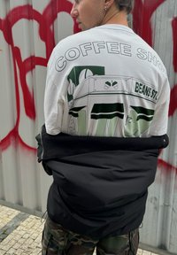T-shirt grafico bianco con illustrazione verde di una caffetteria e il testo "BEANS ST.". Giacca nera in tessuto testurizzato appoggiata sulle spalle.