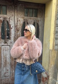 Femeie blondă, purtând o haină roz de blană, fustă din denim și ochelari de soare, ținând o geantă matlasată de culoare bleumarin, stă în fața unor uși din lemn uzat.