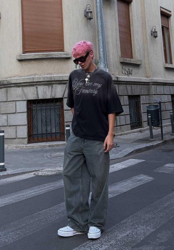 Jeune personne avec des cheveux roses, des lunettes de soleil noires, un t-shirt noir avec texte, un jean gris ample et des baskets blanches debout sur un passage pour piétons urbain.