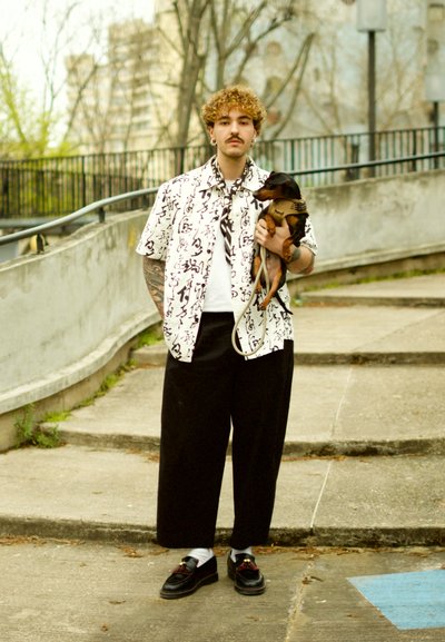 Camisa blanca de manga corta con estampado abstracto negro, pantalones anchos negros, zapatos negros con acentos rojos, sosteniendo un pequeño perro marrón.