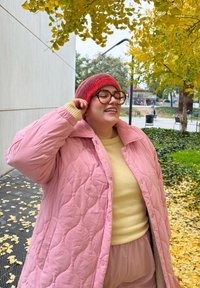 Άτομο που φοράει ροζ καπιτονέ παλτό, κίτρινο πουλόβερ και κόκκινο καπέλο, χαμογελώντας έξω δίπλα σε κίτρινα φθινοπωρινά φύλλα.