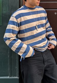 Svītrota garām piedurknēm izstrādājums ar plašām zila un krēma svītrām, kokvilnas audums, krūšu kabata un aproces ar pogām, apvienots ar melniem biksēm.