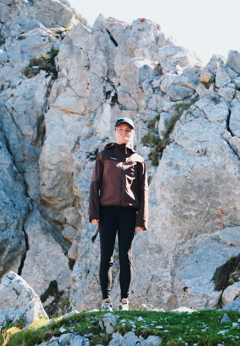 Mladá žena v outdoorové bundě a čepici stojí na travnatém skalnatém terénu s velkými kamenitými útesy v pozadí za jasného denního světla.