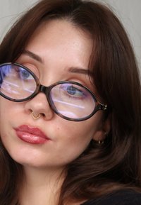 Gafas grandes, redondeadas y de marco oscuro con un brillo translúcido. Un aro de septum dorado acentúa unos labios carnosos con acabado brillante. Piel clara y maquillaje sutil.