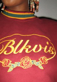Sweatshirt bordeaux com bordados amarelos apresentando o texto "Blkvis" e rosas vermelhas com folhas azul-verde; gola canelada com riscas verdes, amarelas e vermelhas.