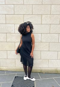 Svart ärmlös klänning med hög halsringning, genomskinligt tygöverdrag, i kombination med vita skor och svarta strumpbyxor, stående mot en stenvägg.