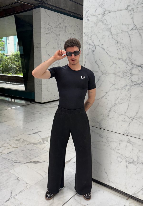 Hombre con camiseta negra de Under Armour, pantalones negros anchos y gafas de sol, de pie en un espacio interior con suelo y paredes de mármol.