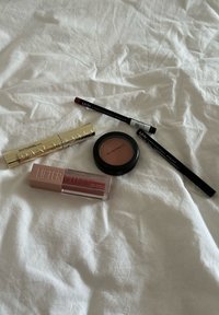 Cinq produits de maquillage sur une surface blanche : un tube de mascara doré, un gloss à lèvres rose, une palette de blush noire et deux crayons eyeliner (noir et rouge).