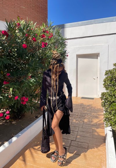 Top negro de manga larga, falda negra con volantes y un corte, cinturón plateado, bolsa de malla negra, sandalias gruesas, rodeada de plantas florecidas.