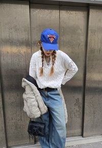 Weißer Kabelstrickpullover, hellblaue Jeans, grauer Kunstfellmantel über einer Schulter, blauer Hut mit orangefarbenem Logo, kleine schwarze Tasche.