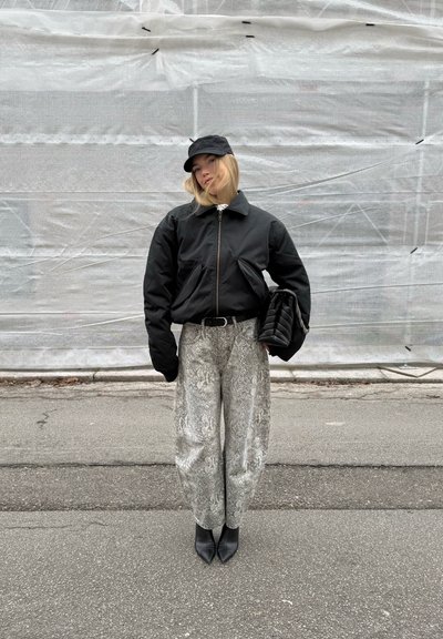 Chaqueta bomber negra con mangas extragrandes, pantalones anchos grises con estampado de serpiente, botas negras de punta puntiaguda y una gorra a juego.