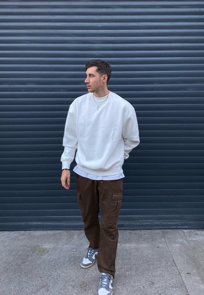 Sudadera blanca con hombros caídos, superpuesta a una camisa blanca; pantalones cargo marrones con bolsillos laterales; zapatillas gris y blancas.