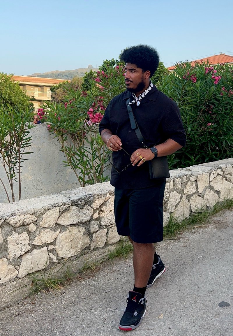 Μαύρο κοντομάνικο πουκάμισο, μαύρες βερμούδες και μαύρα αθλητικά παπούτσια. Τσάντα αξεσουάρ και γυαλιά ηλίου στο χέρι. Το φόντο περιλαμβάνει πράσινα φυτά και έναν πέτρινο τοίχο.