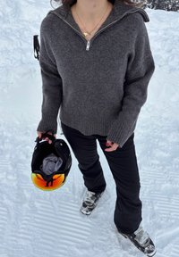 Personne en pull gris et pantalon de ski noir tenant un casque de ski avec une visière orange réfléchissante, debout sur la neige.