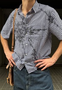 Svart-hvitt gingham skjorte med korte ermer, knapp-down krage og intrikate svarte broderier. Bært med mørke denimjeans og en teksturert veske.