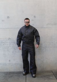 Svart skinnjakke med høy krage, matchende bukser og reflekterende solbriller. Har en teksturert finish og cargolommer på buksene.