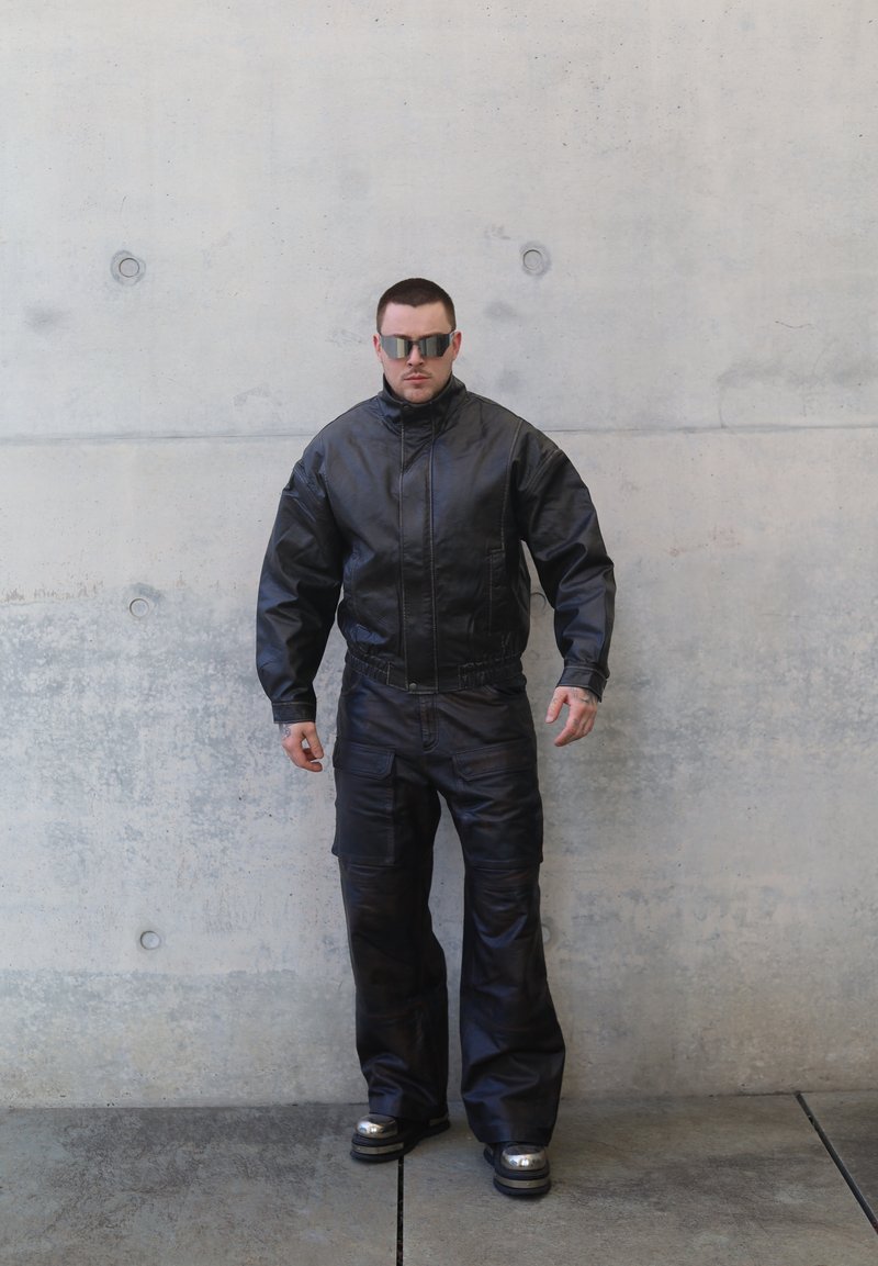 Veste en cuir noire à col montant, pantalon assorti et lunettes de soleil réfléchissantes. Présente une finition texturée et des poches cargo sur le pantalon.
