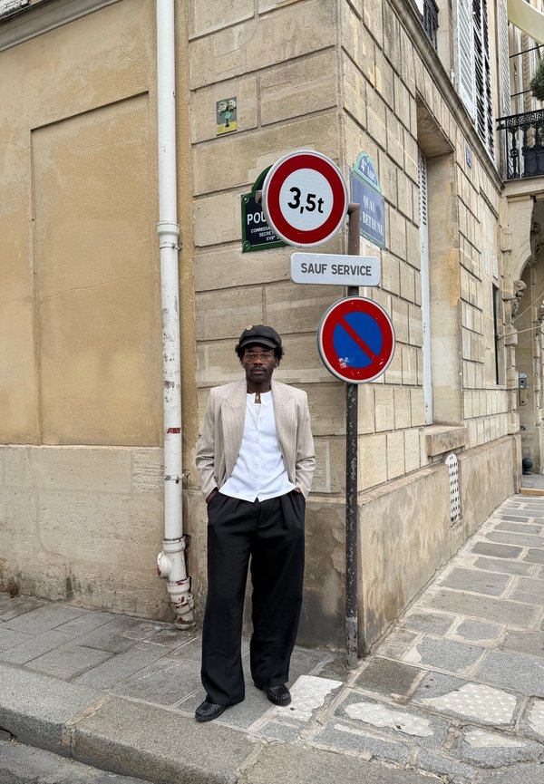 Ein Mann mit leichtem Blazer, weissem Hemd, schwarzer Hose und Hut steht auf dem Trottoir einer Stadt nahe einer Ecke mit Verkehrs- und Parkverbotsschildern.