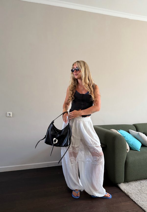 Donna con lunghi capelli biondi che indossa un top nero, pantaloni bianchi in pizzo, sandali blu, occhiali da sole, che tiene una borsa nera in soggiorno.