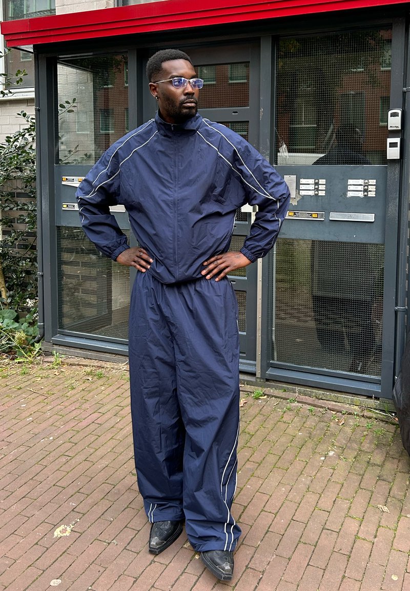 Tuta blu navy con strisce bianche, composto da giacca con zip e pantaloni larghi. Indossato con stivali di pelle neri e occhiali.