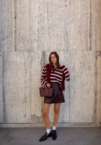 Mujer con suéter a rayas burdeos, falda de cuero y loafers se encuentra de pie contra una pared de piedra texturizada sosteniendo un bolso a juego.
