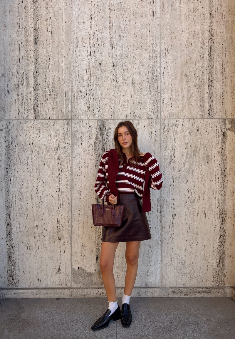 Femme en pull bordeaux à rayures, jupe en cuir et mocassins, se tient devant un mur en pierre texturé en tenant un sac assorti.