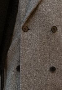 Grijze wollen jas met een dubbele rij knopen, voorzien van vier zwarte knopen en een zachte, gestructureerde stof. Opvallend reversdetail zichtbaar.