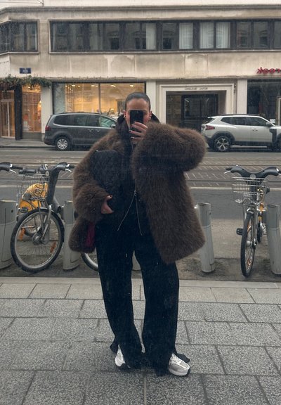 Abrigo grande y esponjoso marrón, chaqueta negra con cremallera, pantalones negros, zapatillas plateadas. Reflejo en un espejo con la calle y bicicletas al fondo.