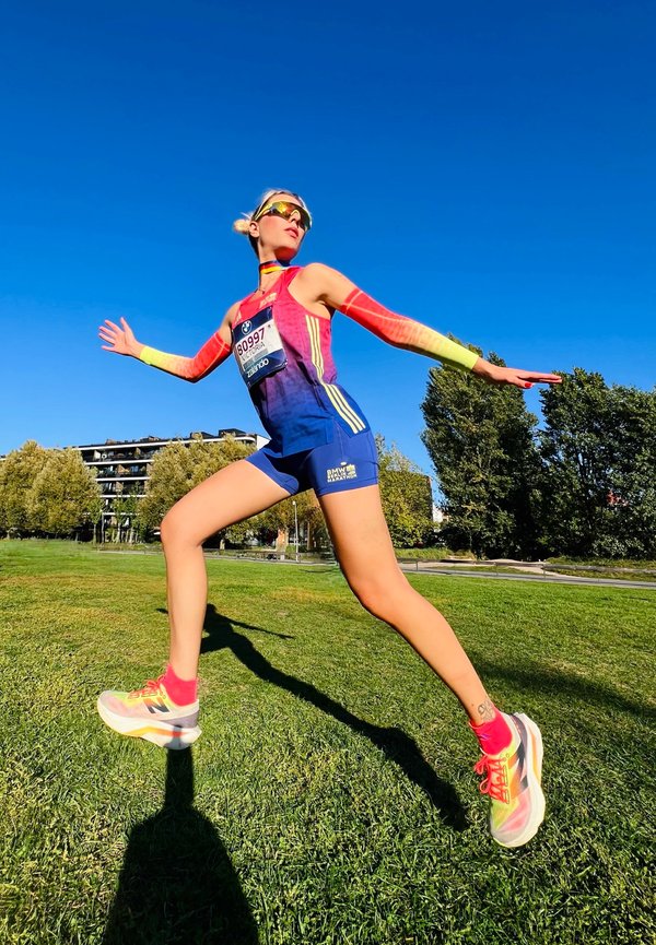 Atleta con ropa de correr colorida salta sobre hierba verde con los brazos extendidos bajo un cielo azul claro cerca de árboles y edificios.