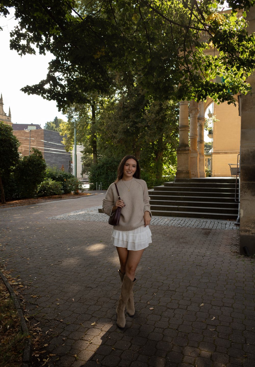 Beige strikket sweater, hvid flæset nederdel og lysebrune knæhøje støvler. Mørk taske. Udendørs omgivelser med træer og stenarkitektur.