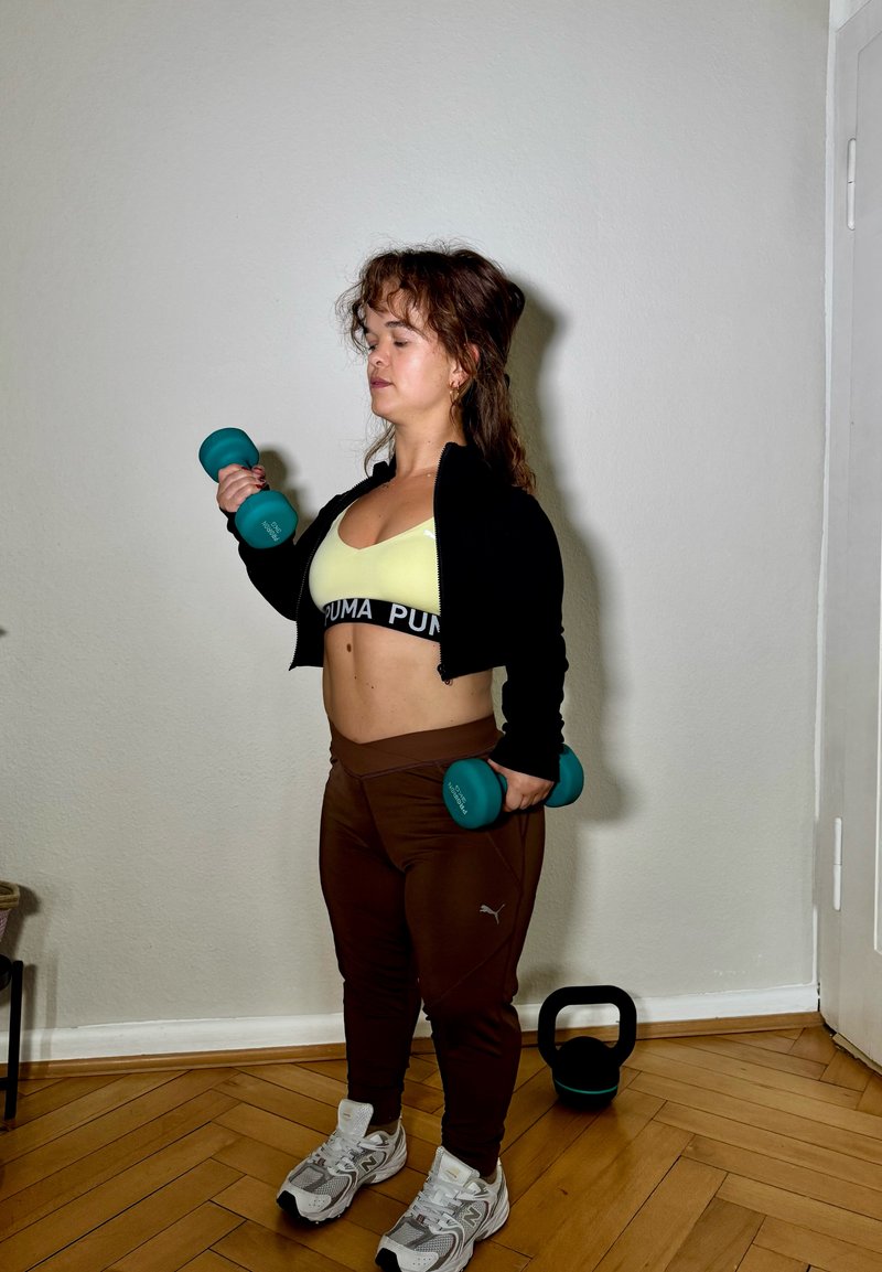 Due kettlebell turchesi tenuti da una persona che indossa una giacca nera corta, un reggiseno sportivo giallo e leggings marroni. Sono visibili delle scarpe da ginnastica bianche.