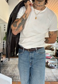 Balta medvilnės marškinėliai, mėlynos džinsinės kelnės, rudas odinis diržas. Rudas odinis švarkas numestas ant peties, demonstruojantis išsamius tatuiruotės ant rankų.