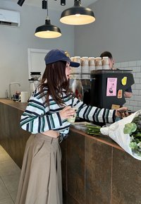 Jeune femme en chemise rayée et casquette payant au comptoir d'un café, tenant une boisson et un téléphone, avec des fleurs emballées sur le comptoir.