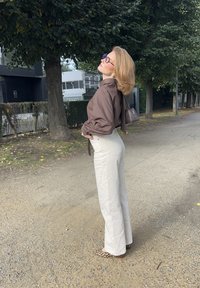 Ruskea pitkähihainen paita, jossa on puffihihat, vaaleankreemiset leveälahkeiset housut ja leopardikuvioiset ballerinat. Pitelee pientä tummanväristä käsilaukkua.