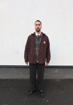 Chaqueta abotonada en tela texturizada roja y negra, encima de un suéter de patrón a cuadros, combinada con pantalones oscuros y zapatos negros gruesos.