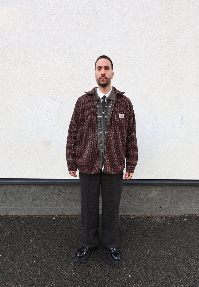 Gombos dzseki piros és fekete texturált anyagból, kockás mintájú pulóver fölött rétegezve, sötét nadrággal és masszív fekete cipővel párosítva.